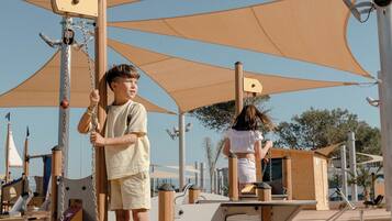 Un parque infantil con columpios y estructuras para escalar, rodeado de arena y cubierto por grandes sombrillas.