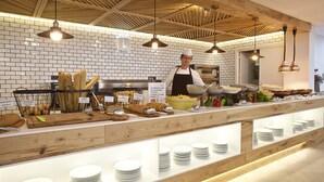 A buffet setup with various food items and utensils, a chef in uniform, and a brick wall background. - Club Hotel Aguamarina Waterpark (Mercadal)