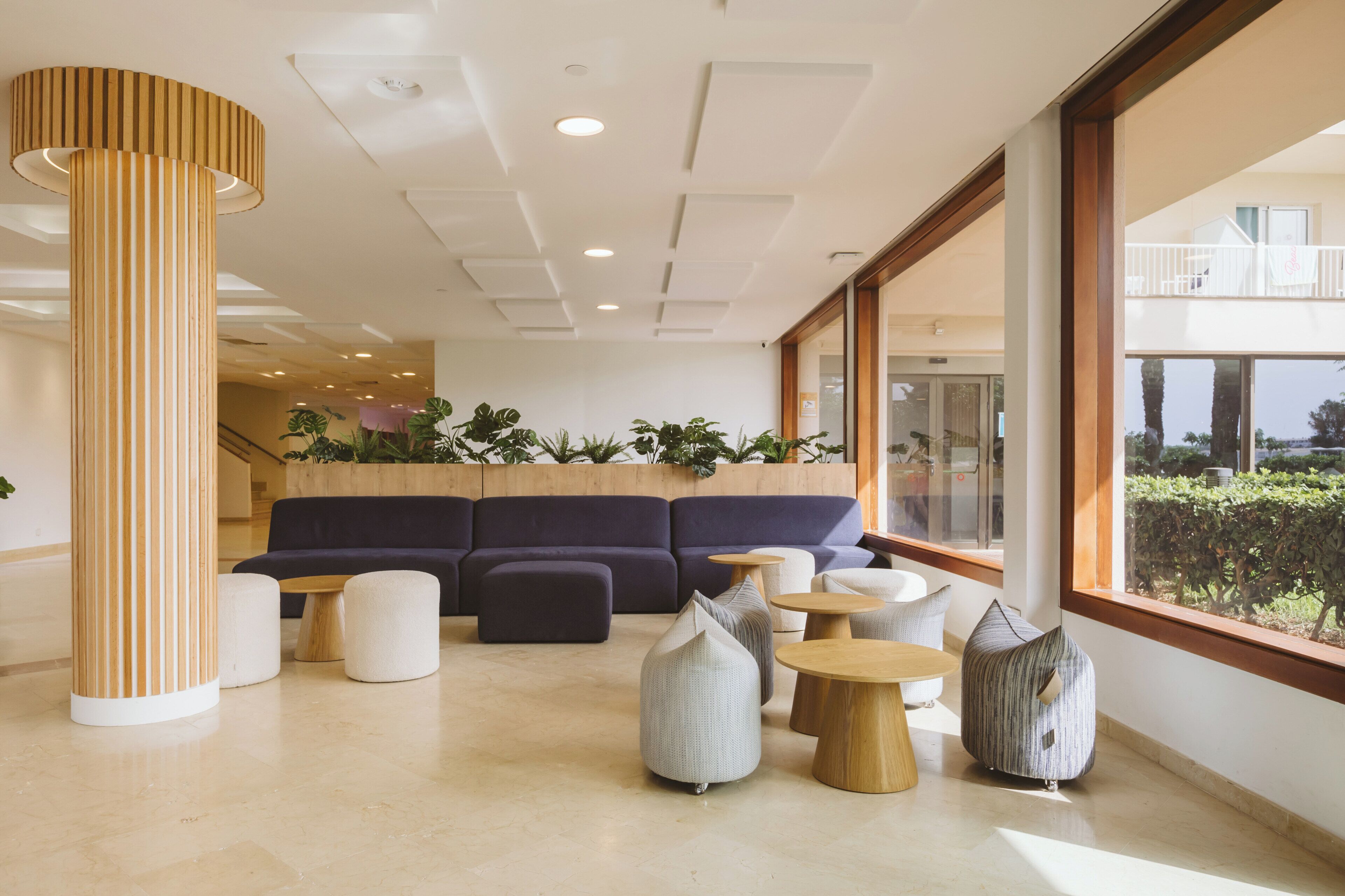 Un espace de détente moderne comprenant un canapé, des poufs et des tables rondes, agrémenté d’une colonne en bois et offrant une vue à travers de grandes fenêtres.