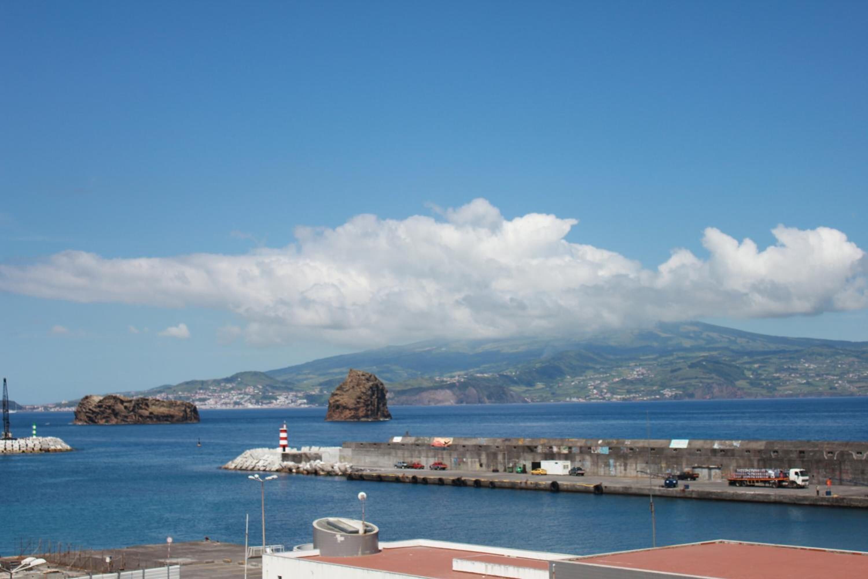 Vista costera con un muelle, afloramientos rocosos y una cadena montañosa distante.
