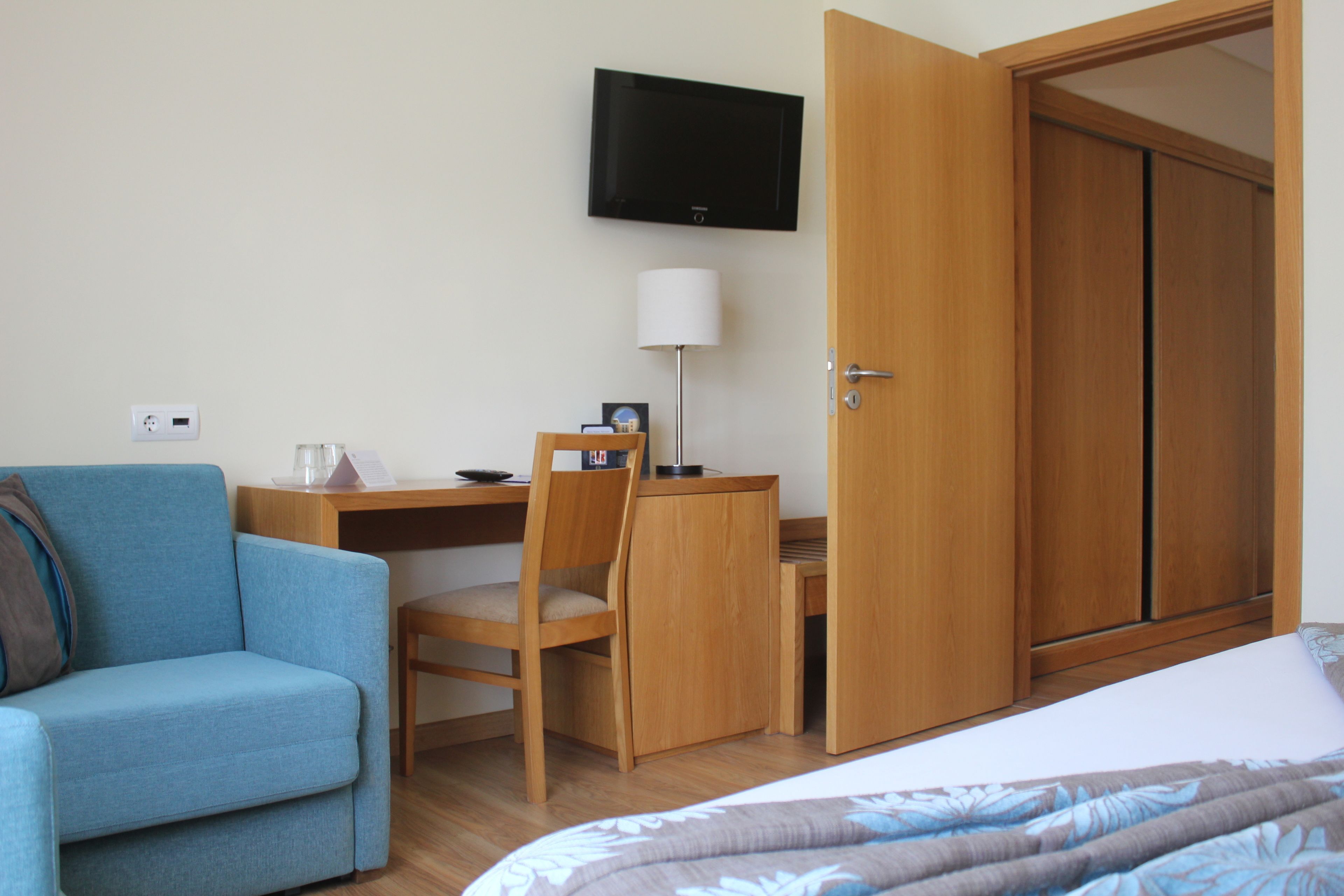 A hotel room with a bed, a desk with a chair, a television mounted on the wall, and a wardrobe.