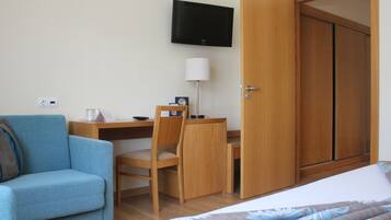 A hotel room with a bed, a desk with a chair, a television mounted on the wall, and a wardrobe.
