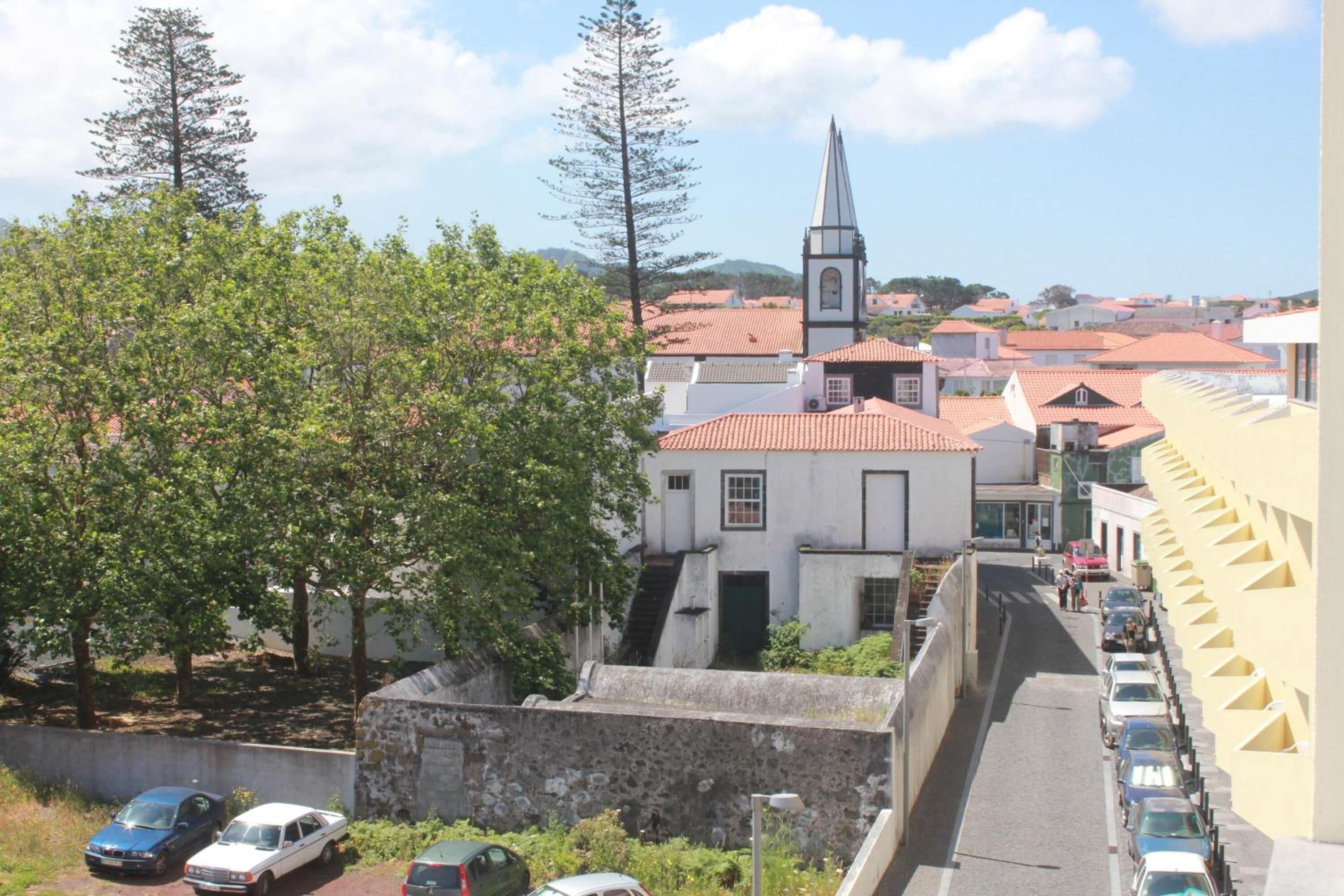 A street view with parked cars, a church with a tall spire, and a stone wall.
