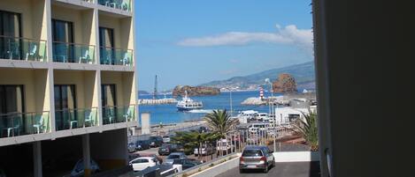 Un hotel de varios pisos con balcones que dan a una carretera costera con coches aparcados y un barco al fondo.