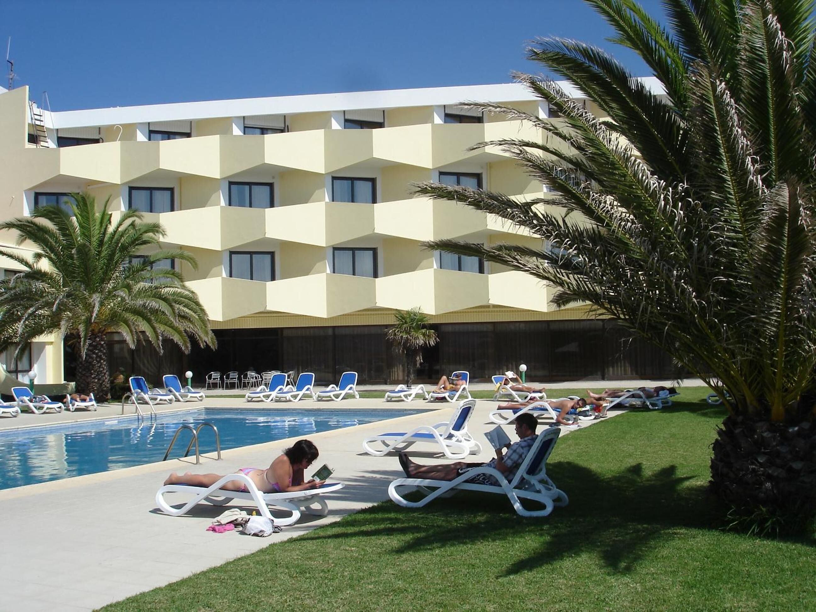 A hotel pool area with multiple lounge chairs and a large building in the background.