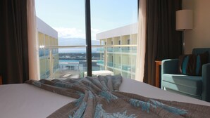 A hotel room with a large bed, a view of a cityscape through floor-to-ceiling windows, a balcony, and a seating area with a lamp. - Hotel Caravelas (Madalena)