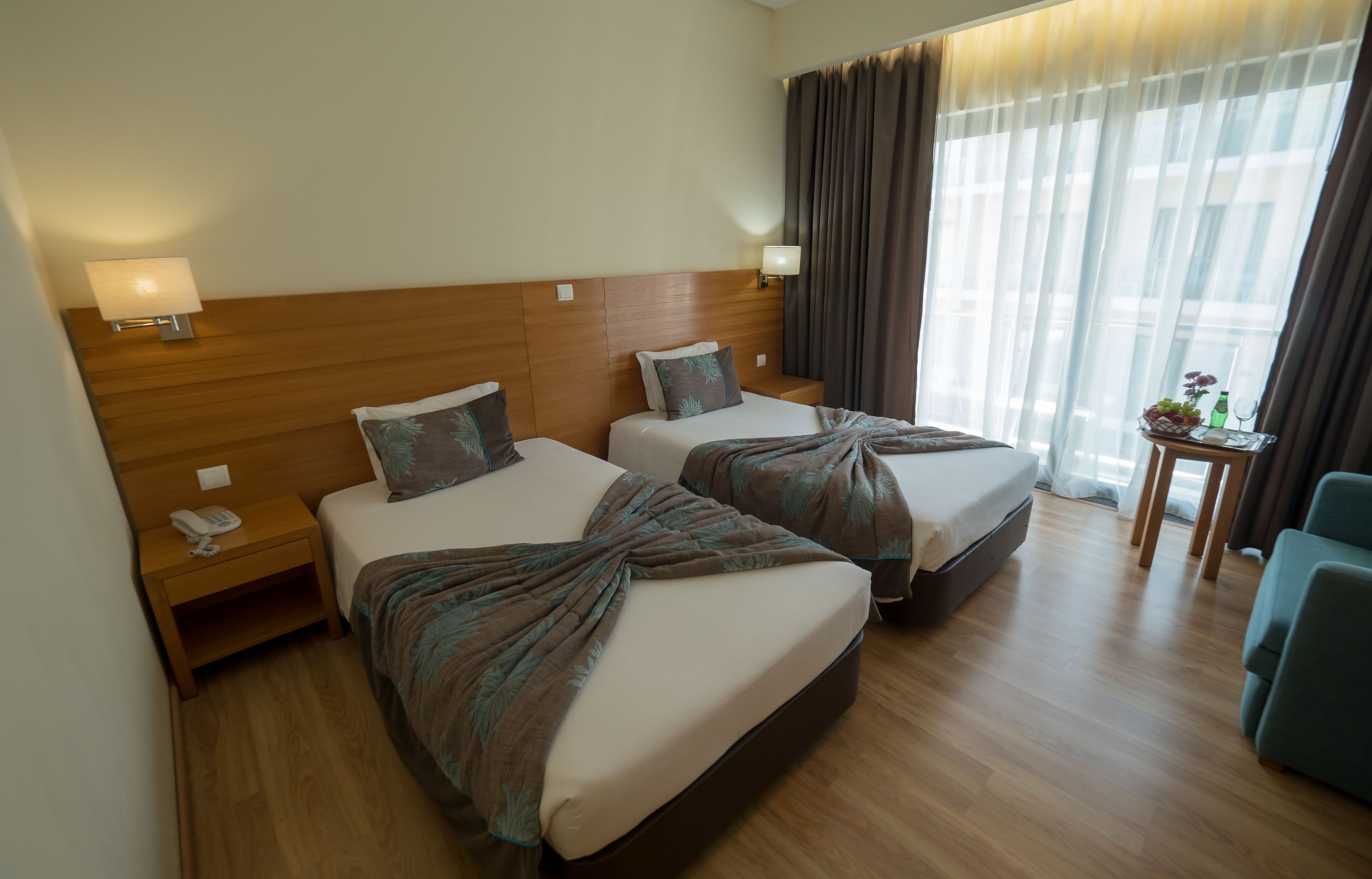 Une chambre d’hôtel avec deux lits, des têtes de lit en bois, des lampes de chevet, une petite table avec un vase, et une vue sur l’extérieur grâce à de grandes fenêtres.
