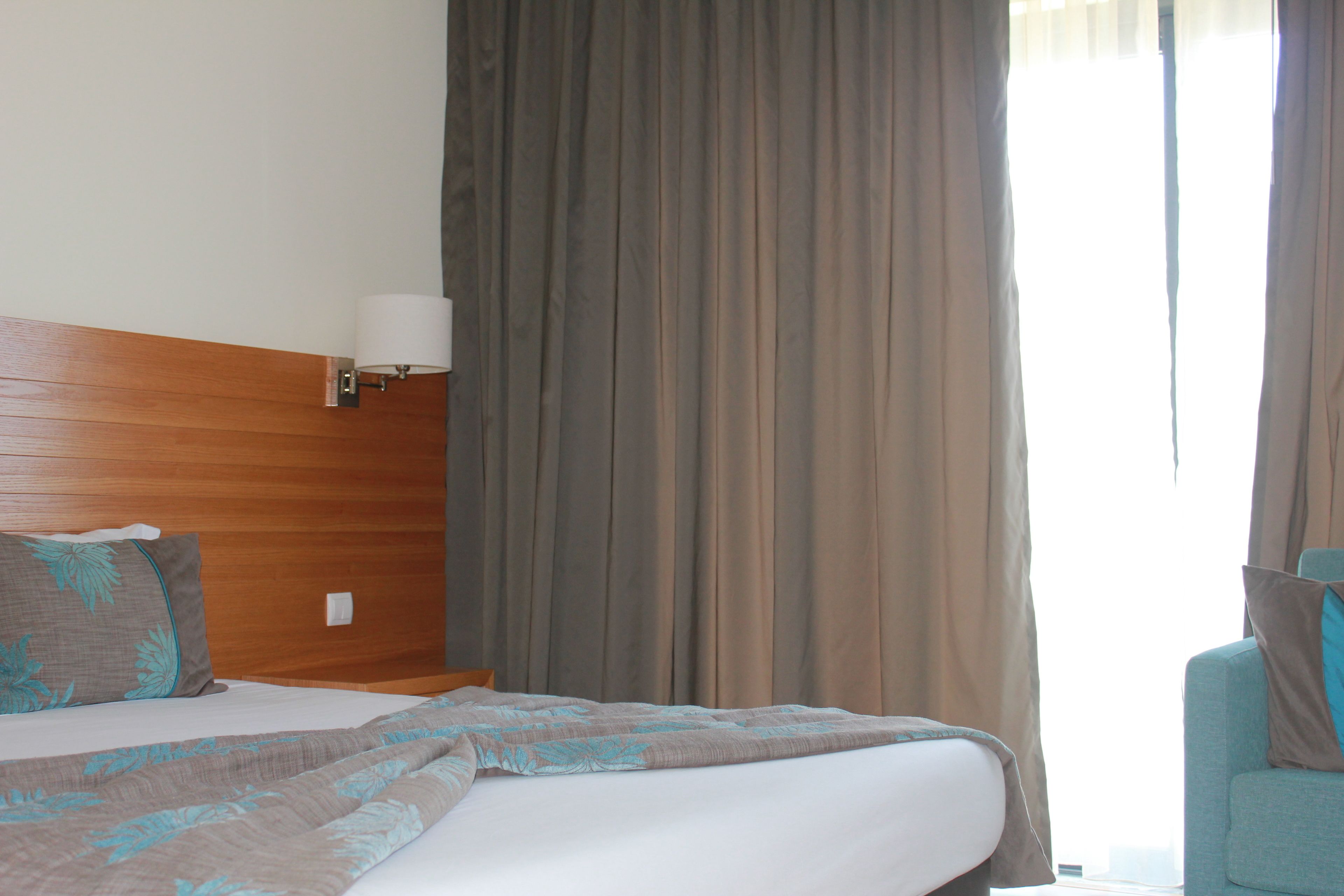 Hotellihuone, jossa on puinen sängynpääty, kuviollinen peitto sängyllä, yöpöytälamppu ja näkymä ulos ikkunasta verhojen läpi.