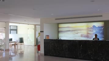 A hotel reception area with a large digital screen displaying a scenic image, a marble reception desk, and a glass partition separating the reception from the adjacent room area.