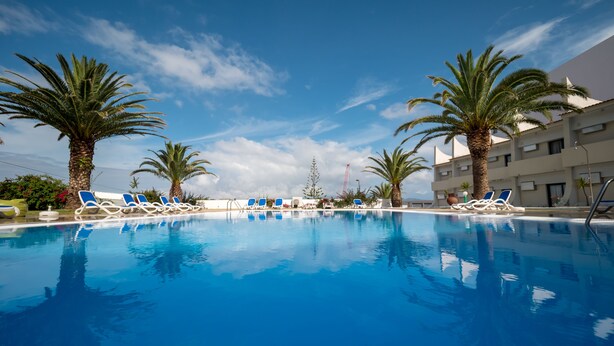 A swimming pool with blue water, surrounded by palm trees and blue lounge chairs. - Hotel Caravelas (Madalena)