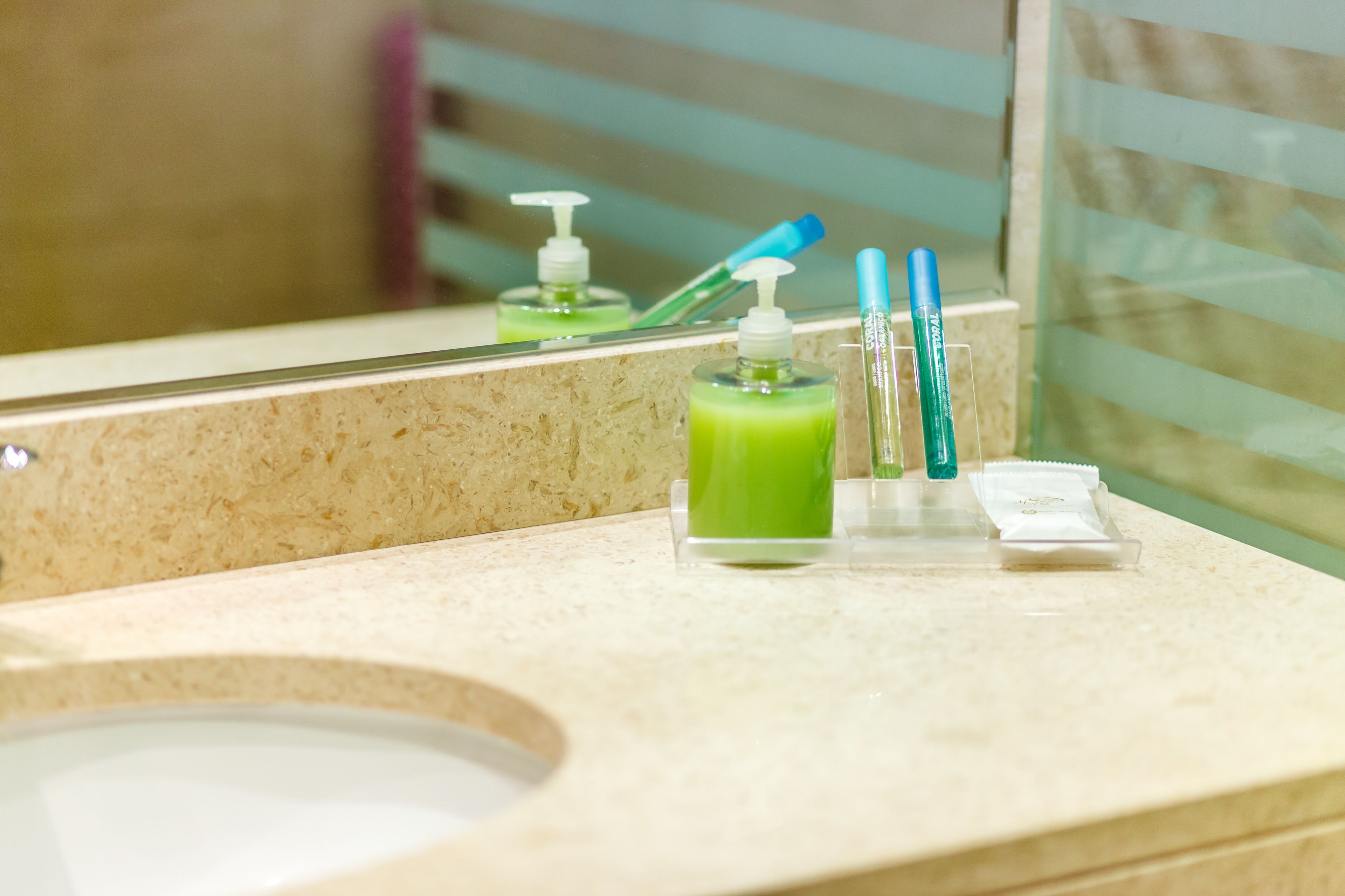 A bathroom with a sink, a mirror, and toiletries including a green bottle, toothbrushes, and a soap dispenser.