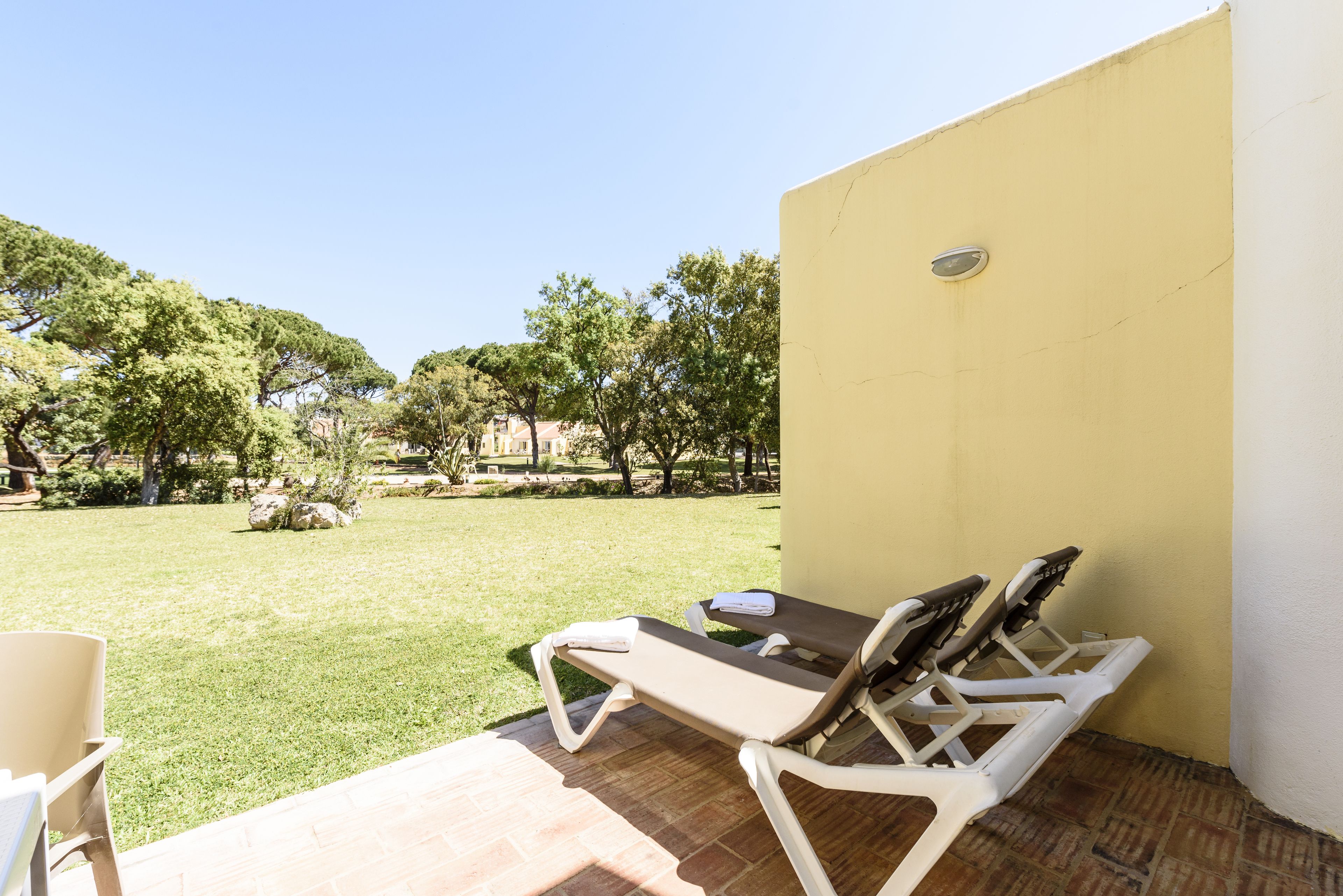 A patio with two sun loungers and a chair, a yellow wall, and a grassy area with trees.