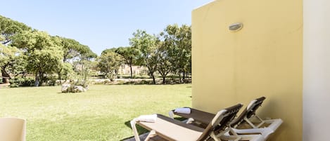 A patio with two sun loungers and a chair, a yellow wall, and a grassy area with trees.