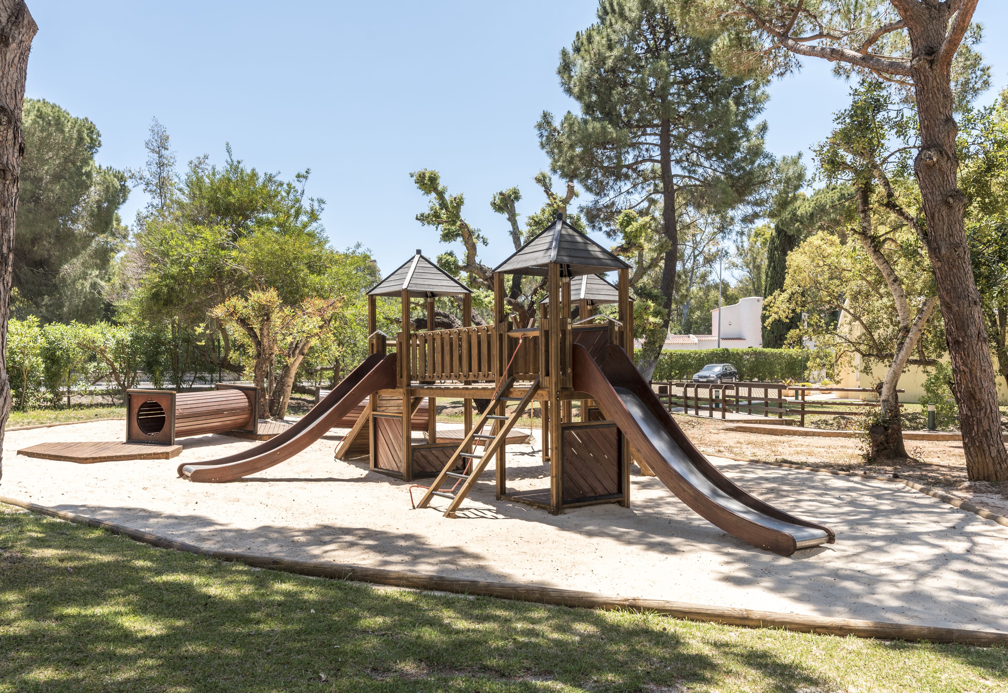A playground with a wooden slide and climbing structures, surrounded by trees and a clear sky.
