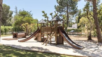 A playground with a wooden slide and climbing structures, surrounded by trees and a clear sky.