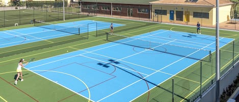 An outdoor tennis court with multiple courts, a fence, and a building in the background.