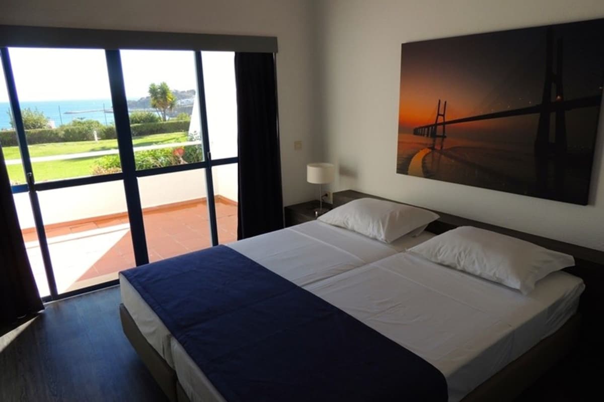 A hotel room with a large bed, a view of the sea through a sliding door, and a painting on the wall.