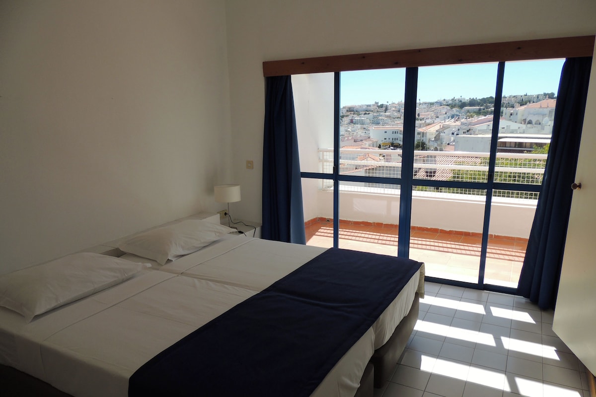 A hotel room with a large bed, a window with curtains, and a view of a cityscape.