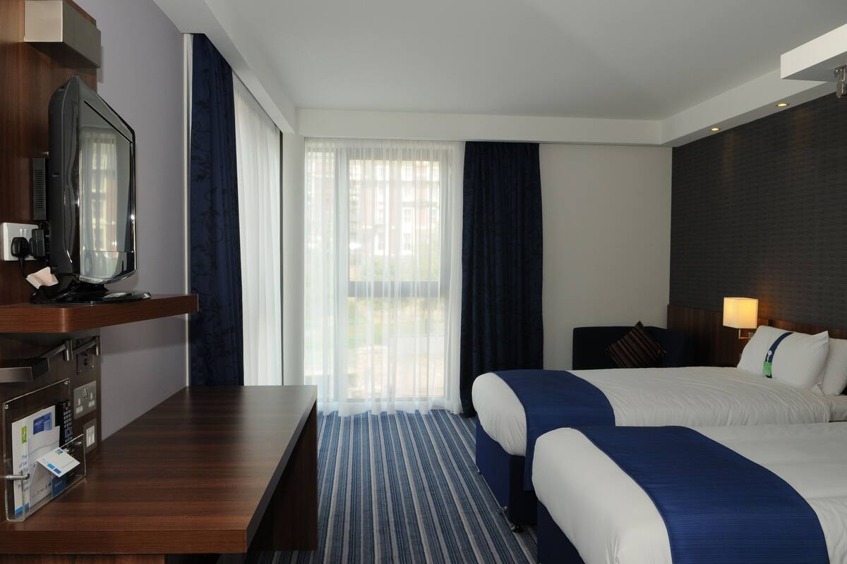 A hotel room with two beds, a desk with a television, a lamp, and a window with curtains.