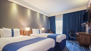 Een hotelkamer met twee bedden, een bureau, een stoel, een televisie en een groot raam met gordijnen.