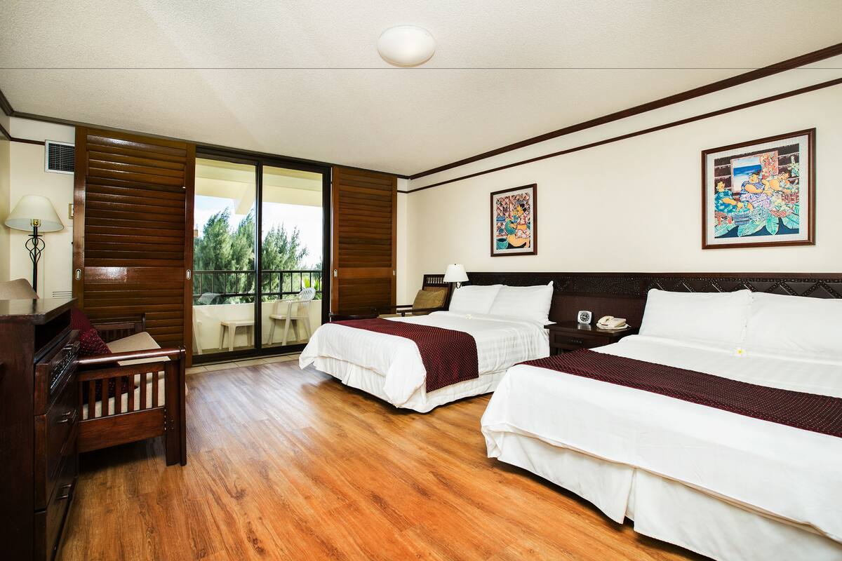 A hotel room with two beds, a wooden desk, a chair, a lamp, and a balcony with a view of trees.