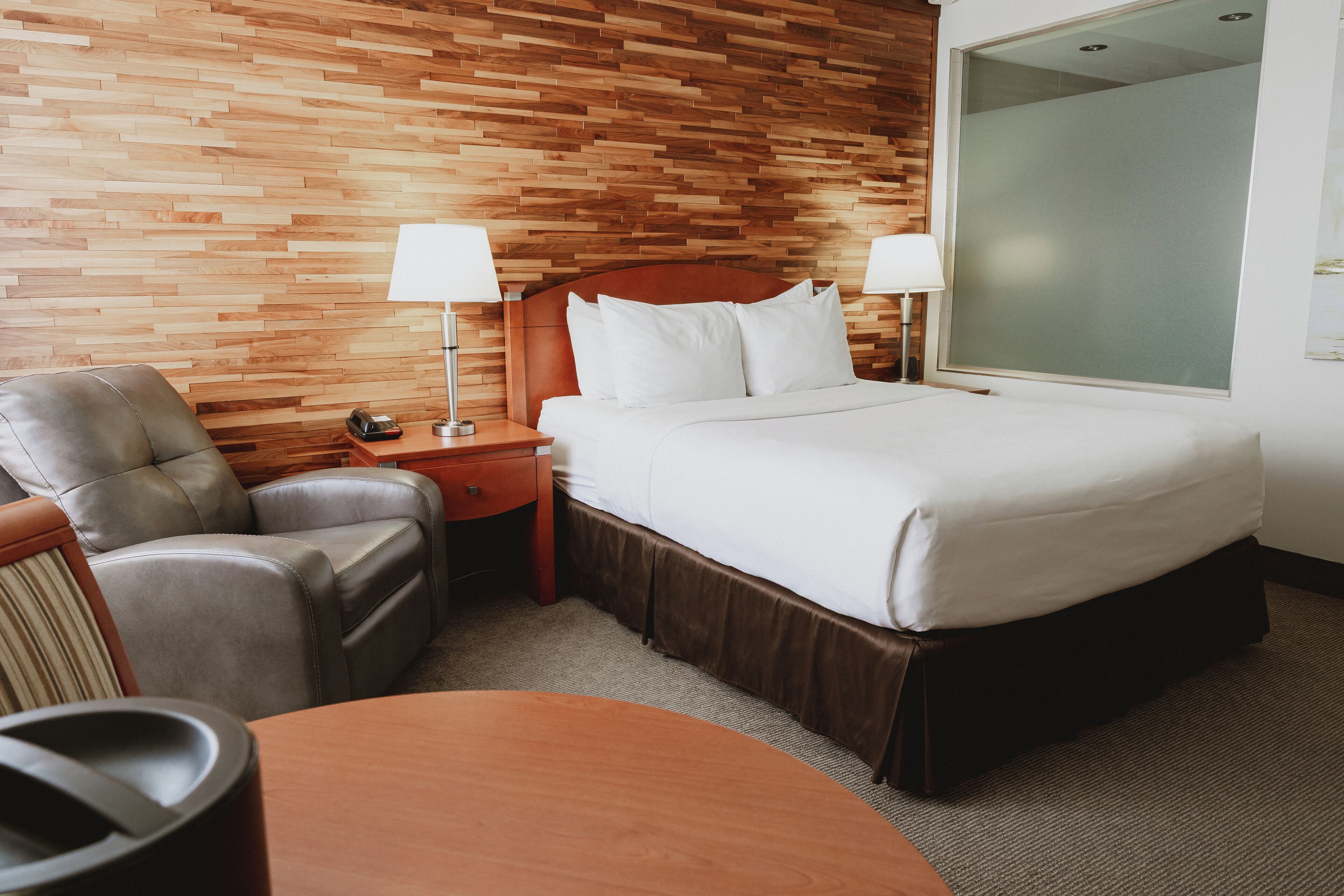 A hotel room with a wooden wall, a bed with white bedding, a leather armchair, a wooden nightstand with a lamp, and a large window.