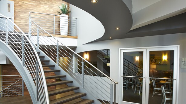 Un interior moderno con una escalera curva, suelo de madera y un área de descanso con sillas y mesas.