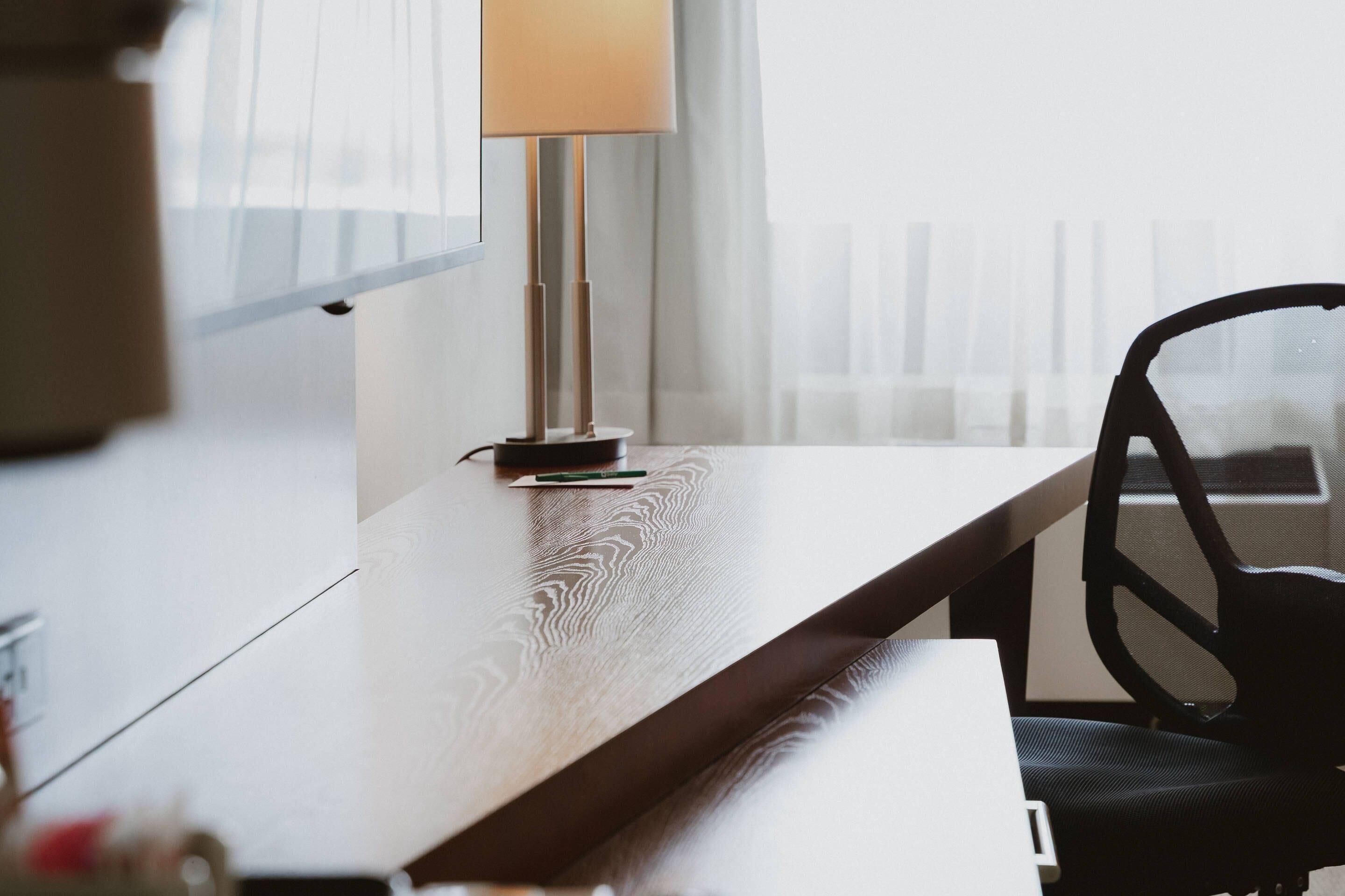 A modern room with a wooden desk, a black office chair, a lamp, and a window with blinds.