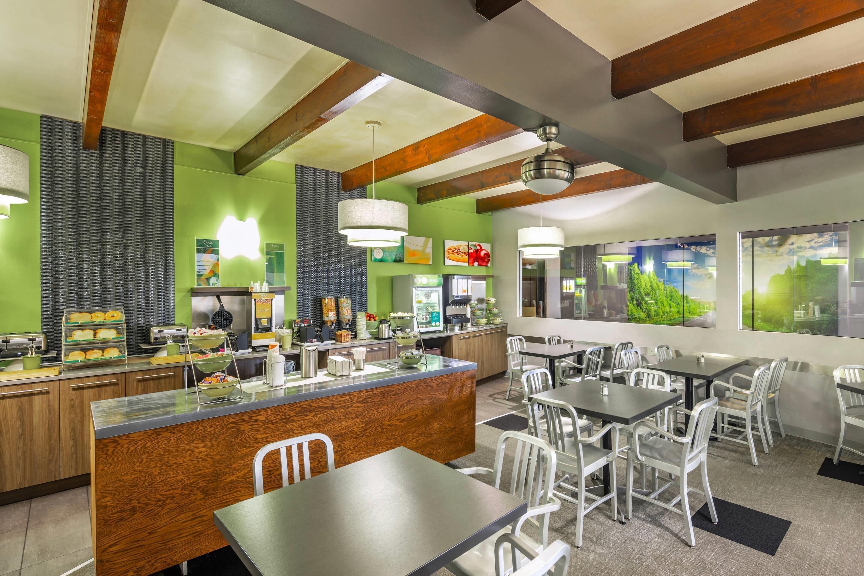 A restaurant with a wooden counter, white chairs, and a view of greenery through large windows.