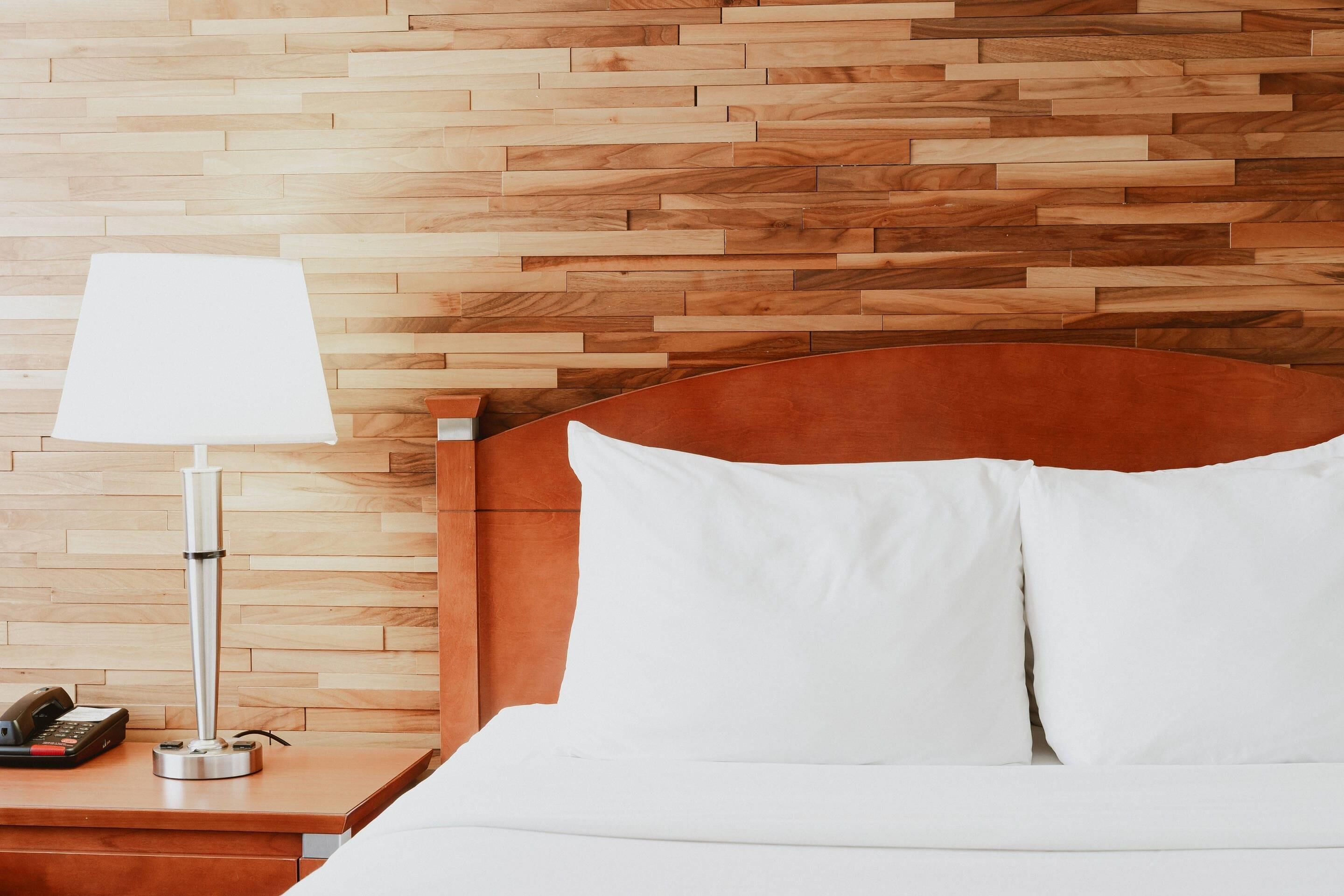A hotel room with a wooden headboard, a bedside table with a lamp, and a bed with white bedding.