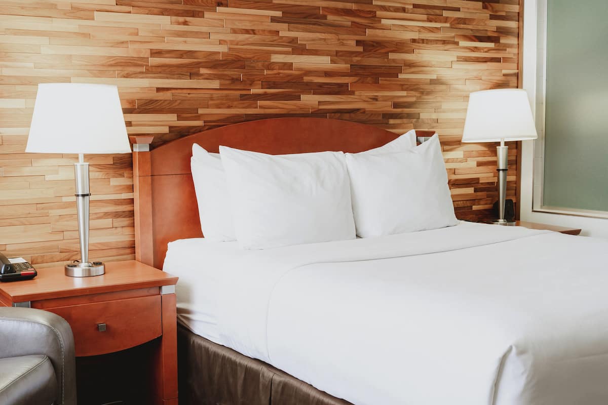 Habitación de hotel con cabecera de madera, dos lámparas de noche, cama con ropa blanca, mesita de noche con teléfono y un cajón.