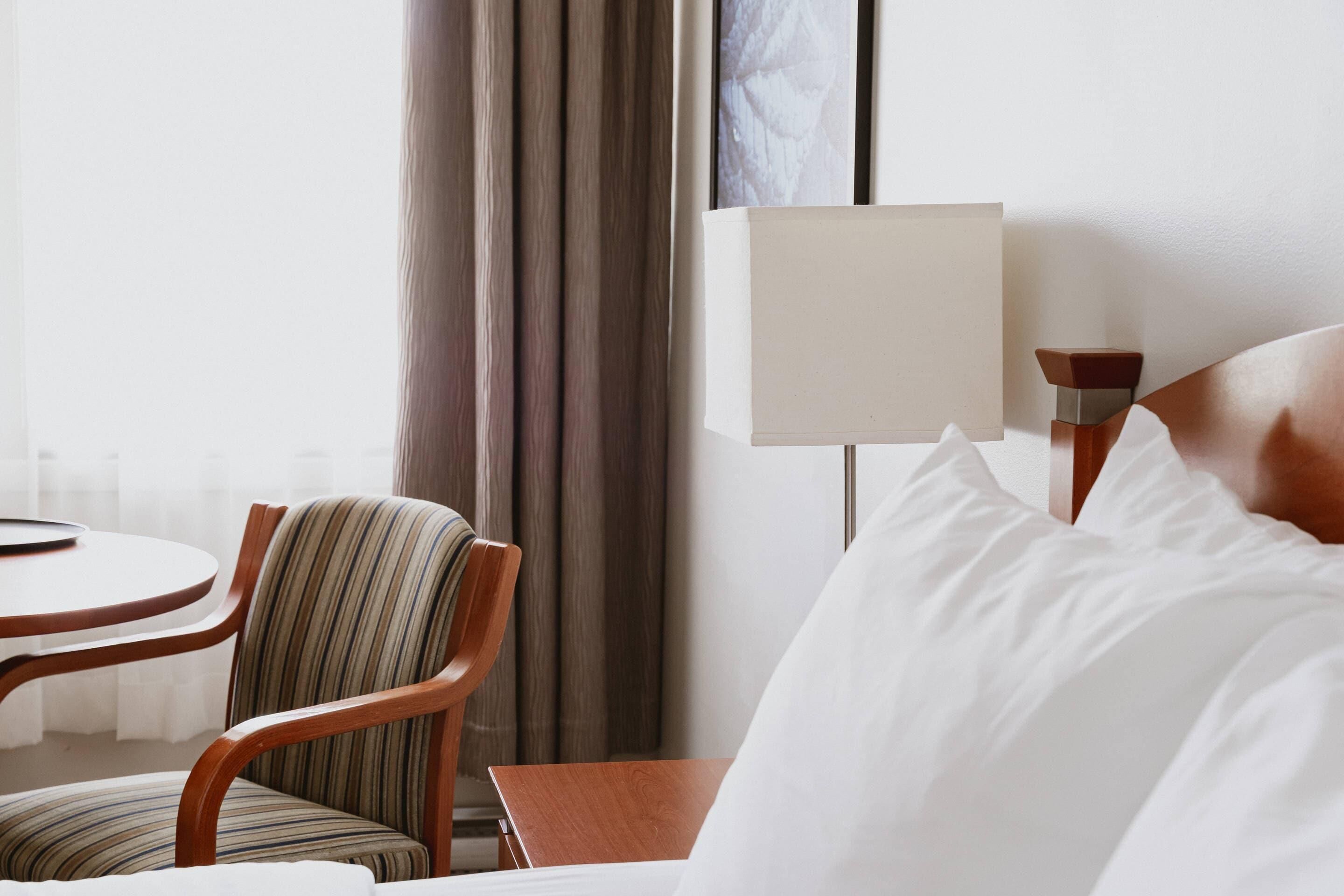 Una habitación de hotel con cama, mesita de noche, lámpara, silla y una pequeña mesa.