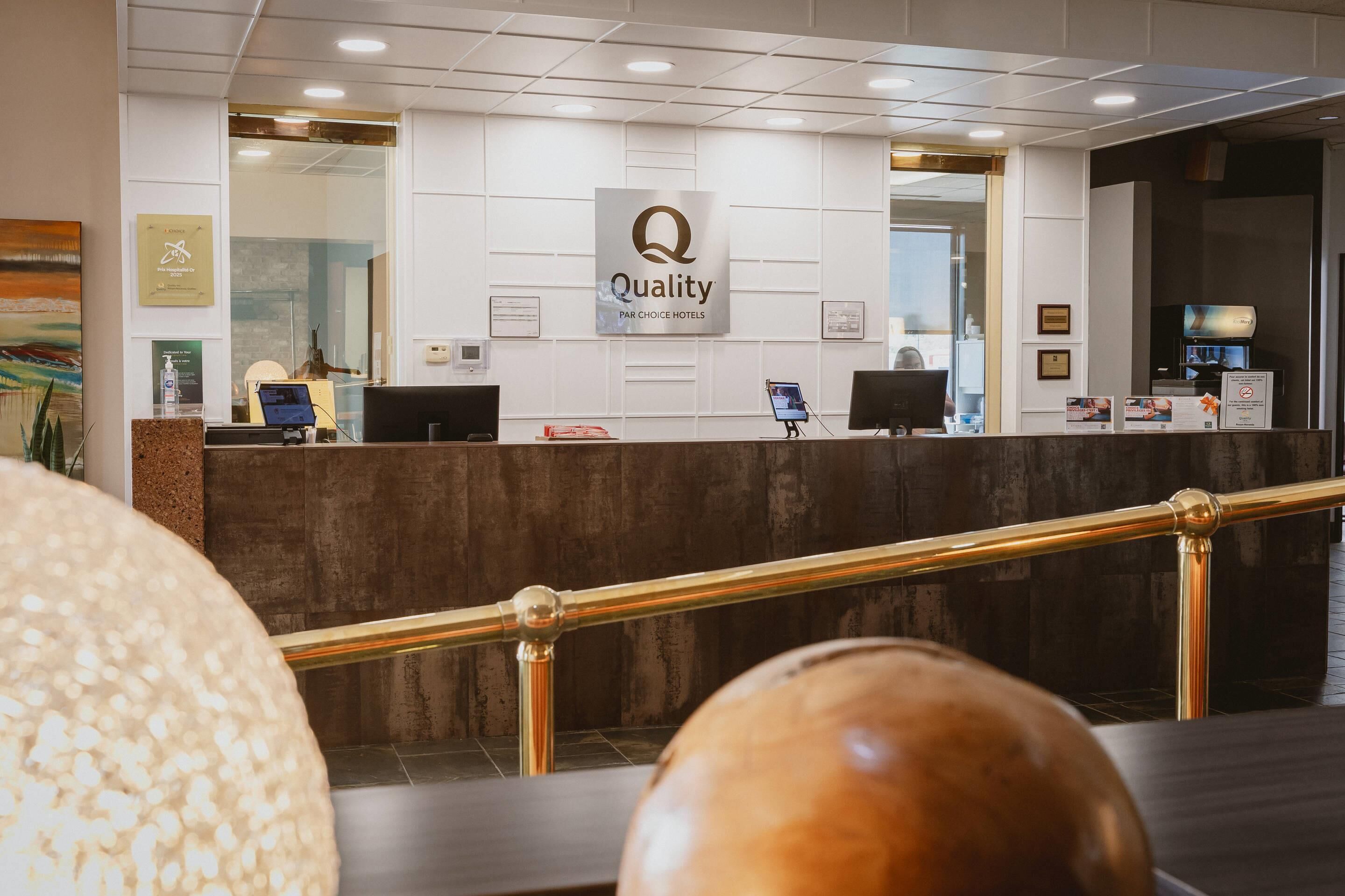 Ein Hotelrezeptionstresen mit einem 'Quality'-Schild, ein Empfangstresen, ein Fernseher und ein gerahmtes Zertifikat an der Wand.