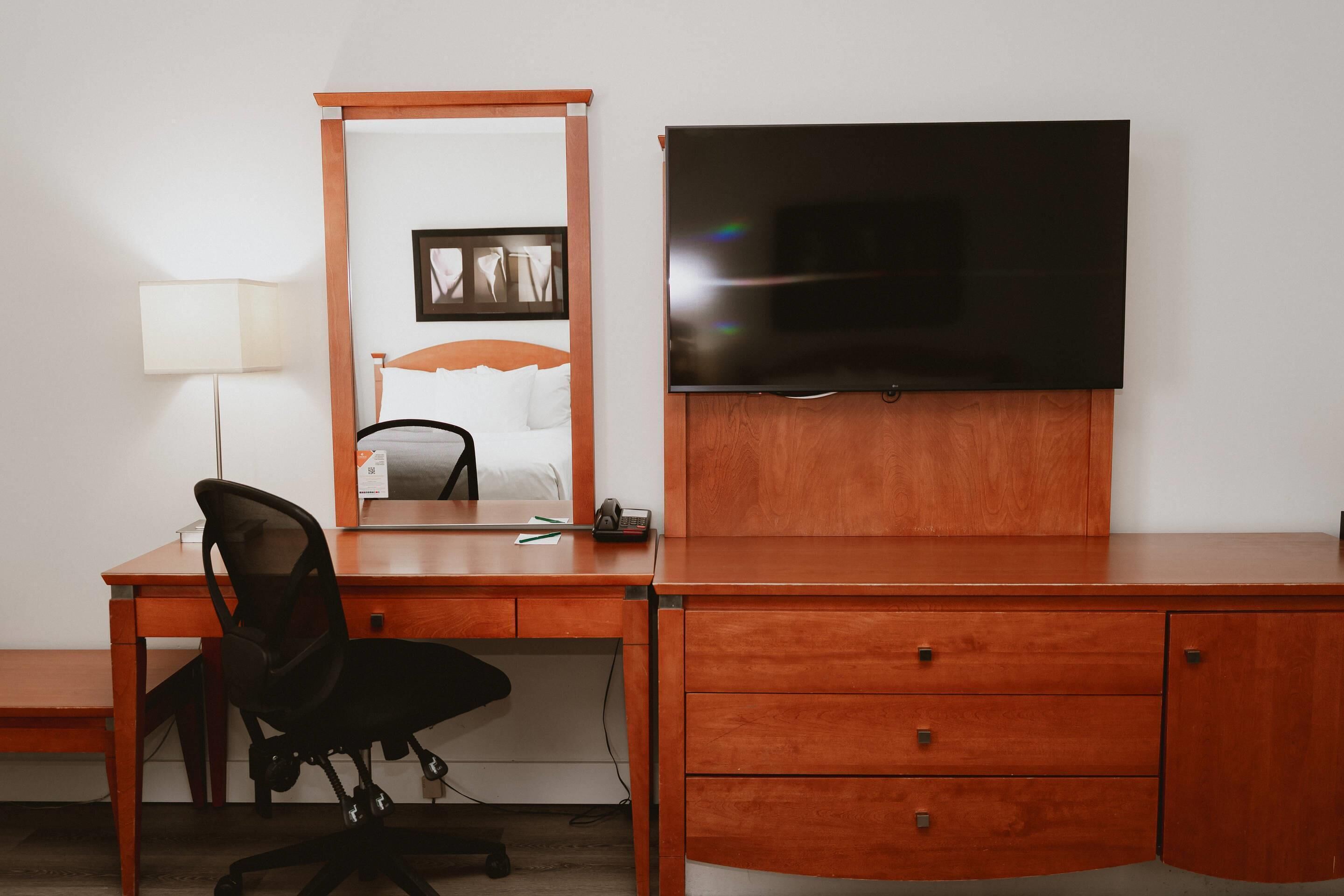 A hotel room with a wooden desk, a black office chair, a flat-screen TV mounted on the wall, a bedside table, and a lamp.