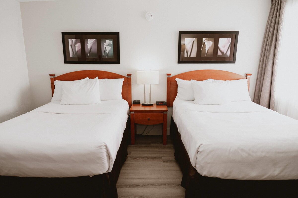 A hotel room with two single beds, a nightstand with a lamp, and framed artwork on the wall.