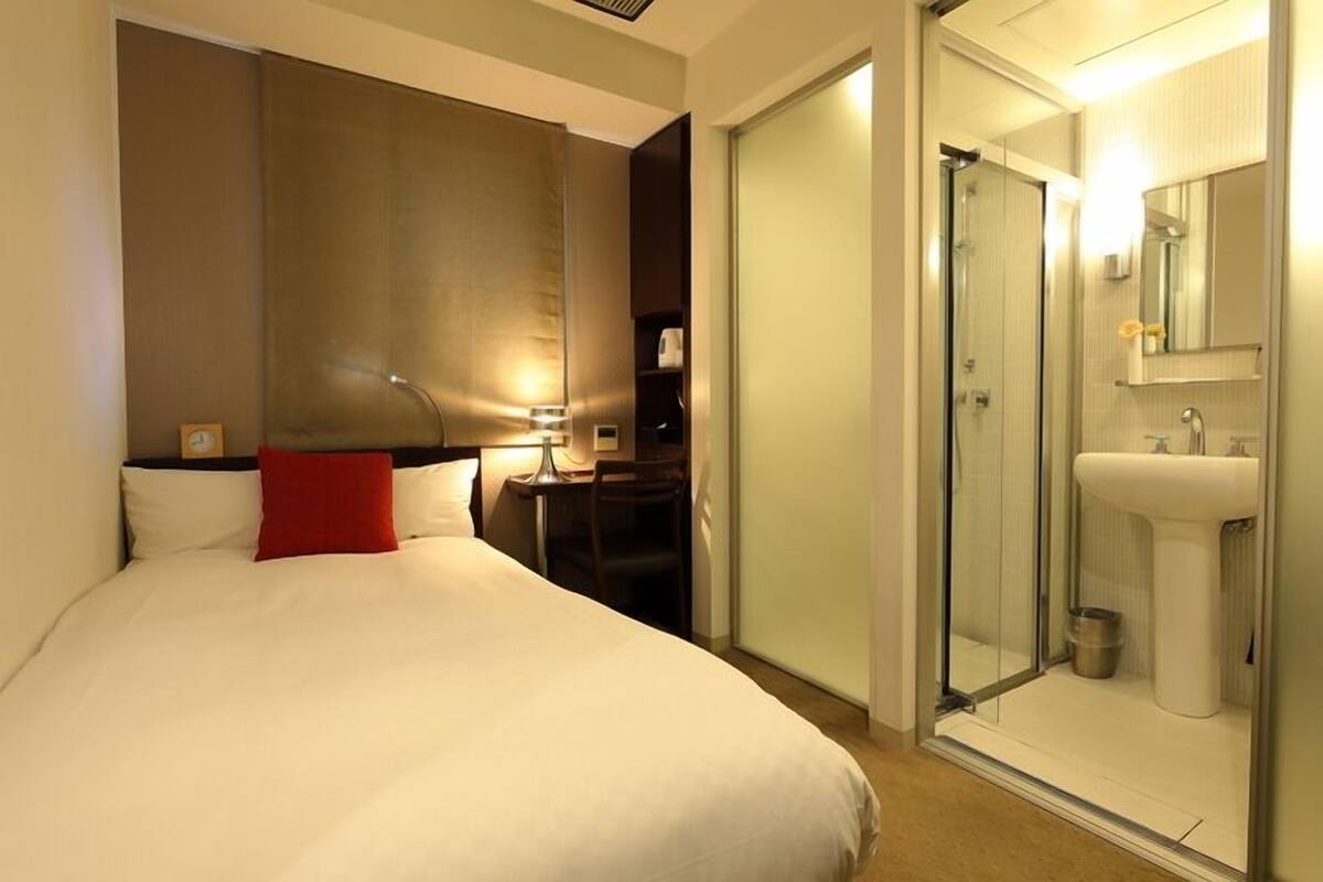 Habitación de hotel con cama, escritorio, silla, mesita de noche, lámpara, cabina de ducha de vidrio y baño con lavamanos y espejo.