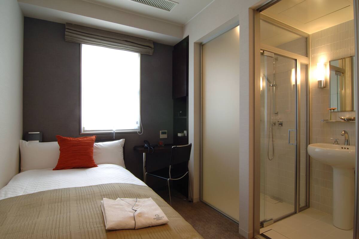 Habitación de hotel con cama, escritorio, silla, zona de ducha y baño con lavamanos.
