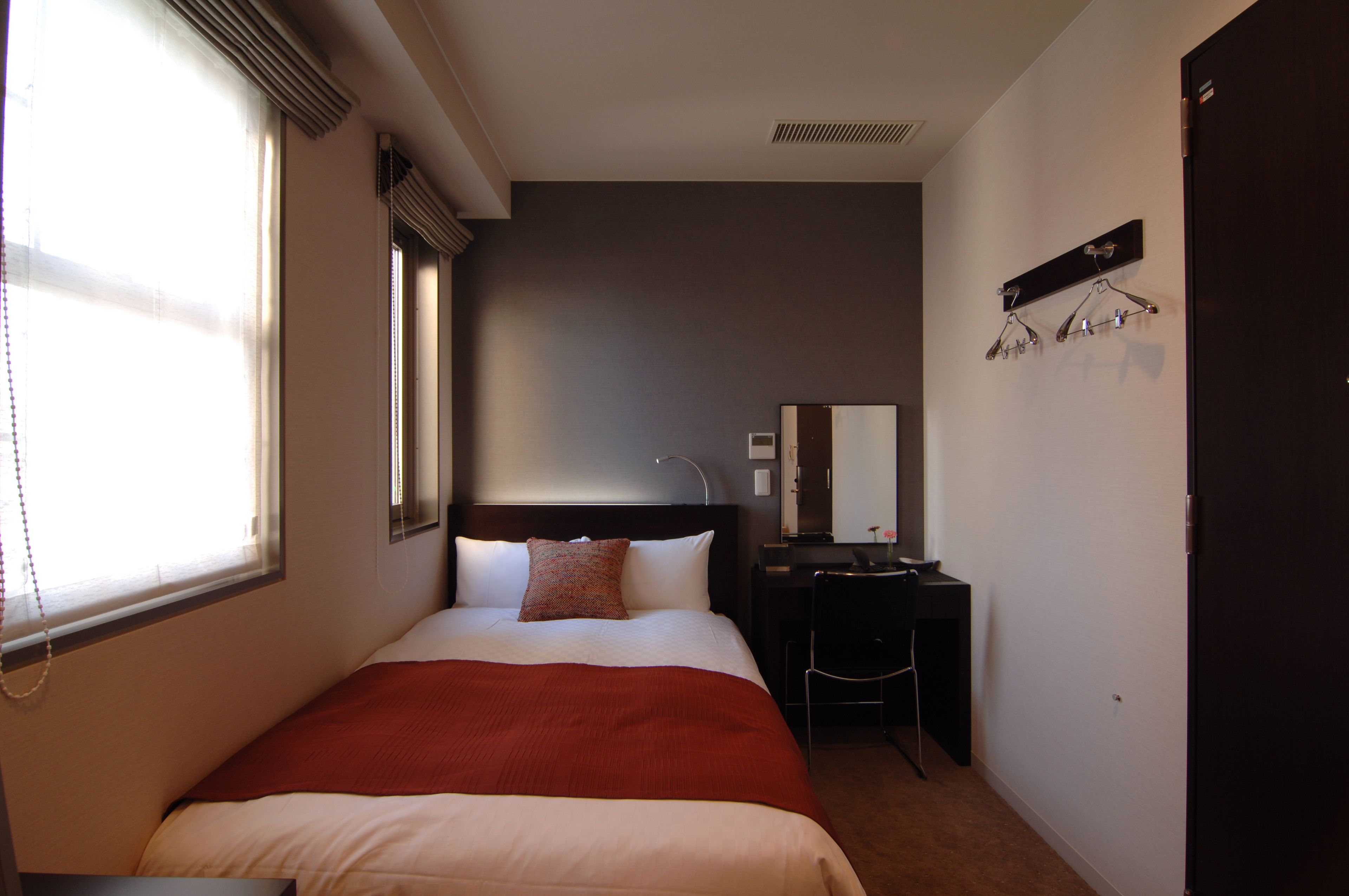 A hotel room with a bed, a desk, a chair, a mirror, and a coat rack.