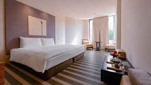 Ein modernes Hotelzimmer mit einem großen Bett, einer kleinen Sitzecke, einem Couchtisch mit Frühstücksangeboten und Blick auf Grün durch das Fenster.