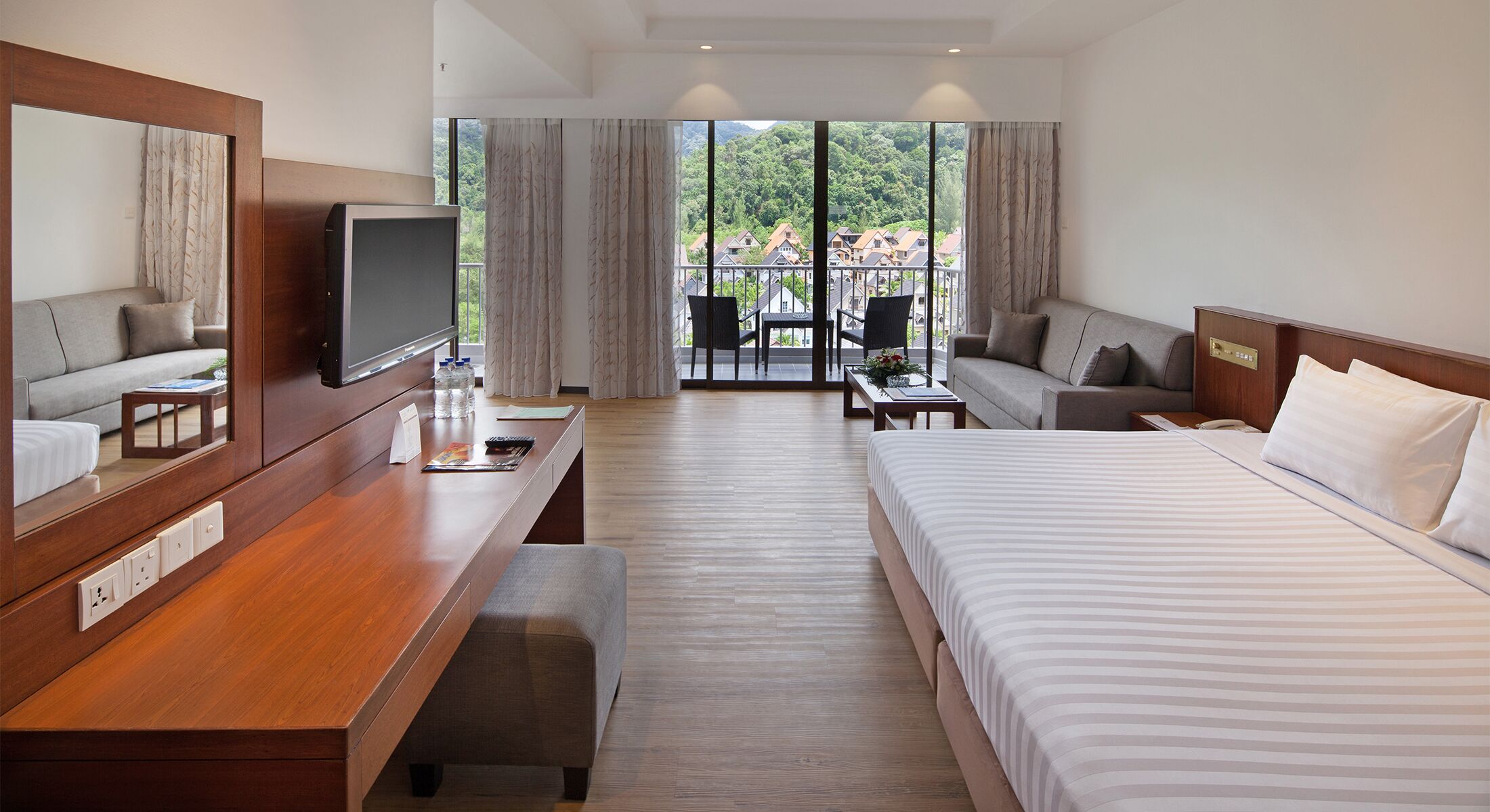 A hotel room with a bed, a desk with a TV, a sofa, and a balcony with a view of houses and trees.