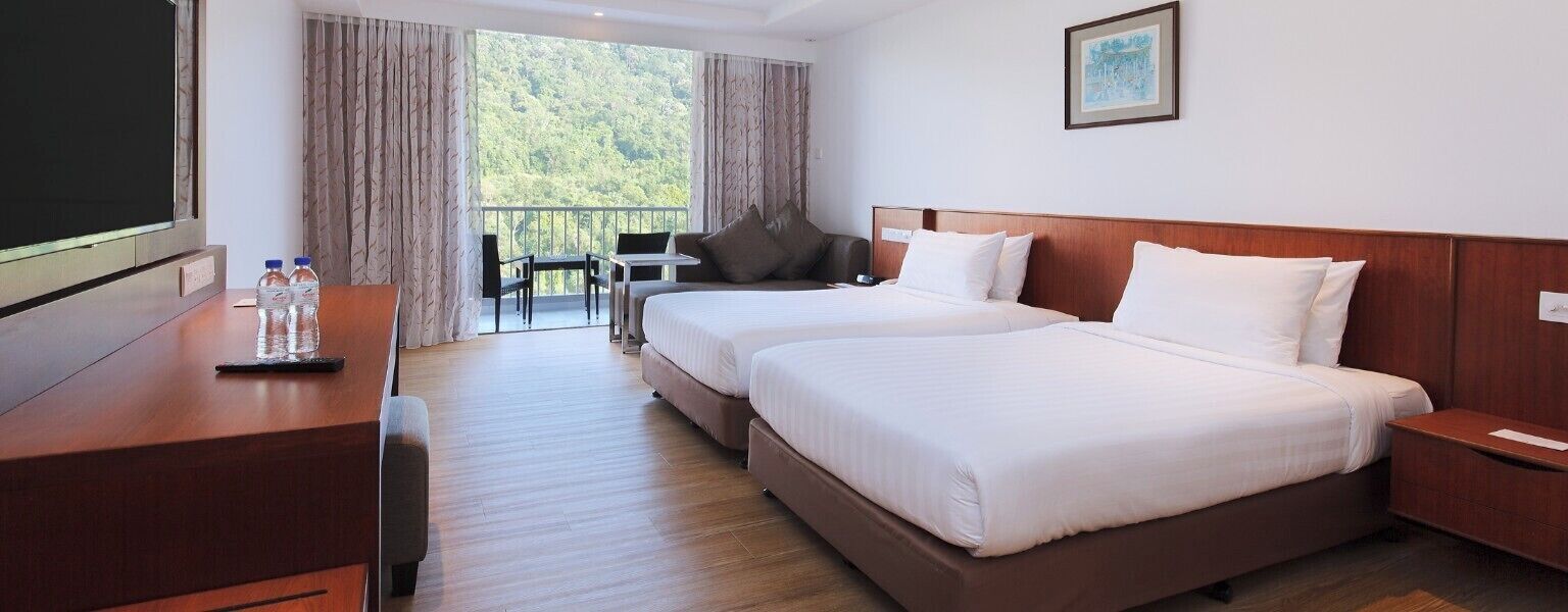 Habitación de hotel con dos camas, un escritorio, un televisor y un balcón con vistas a árboles.