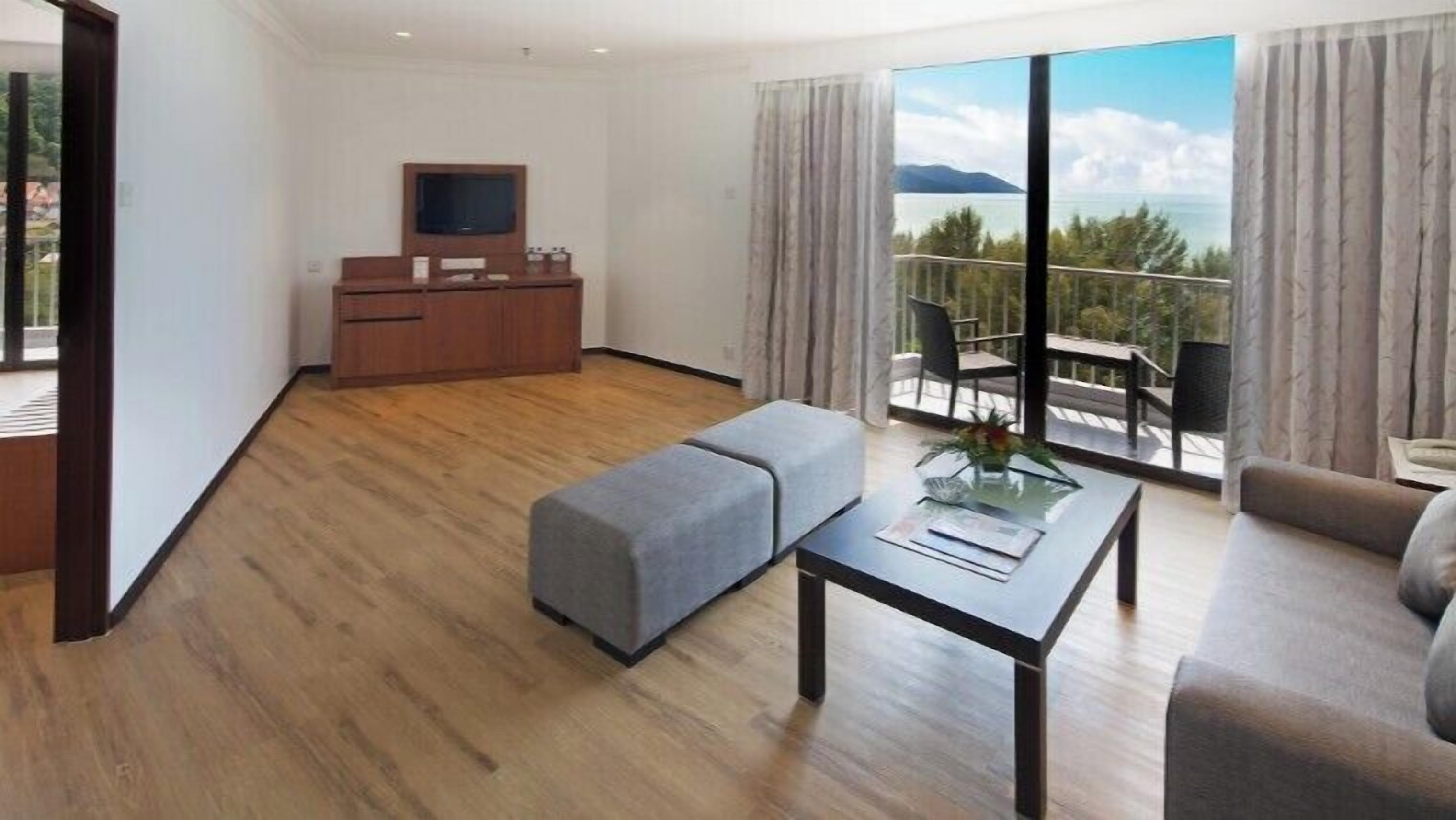 A modern hotel room with a wooden floor, a sofa, a table, a TV, a balcony with a view of the sea, and a kitchenette.