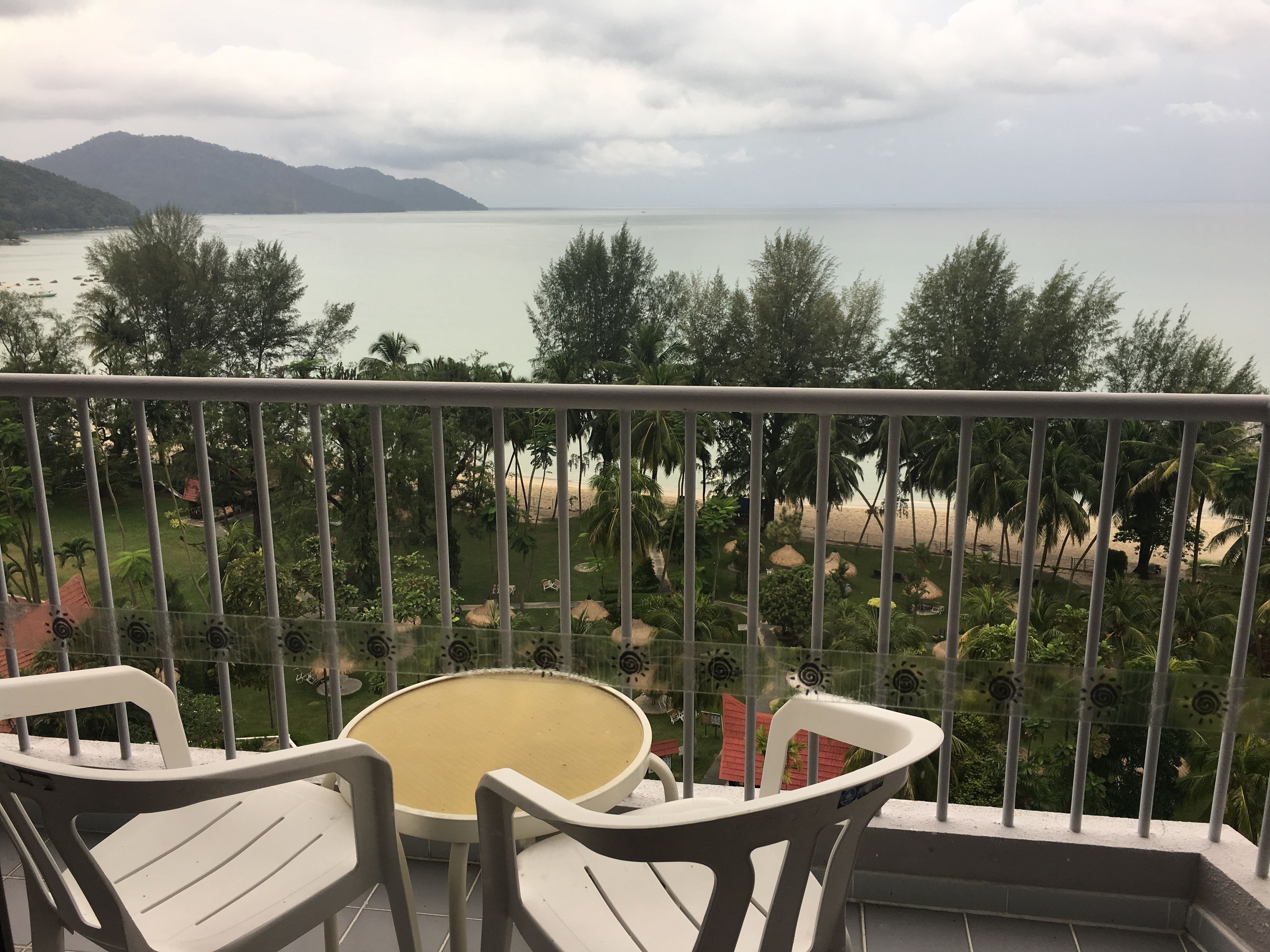 A balcony with a view of a beach and mountains. There are two white plastic chairs and a small round table.