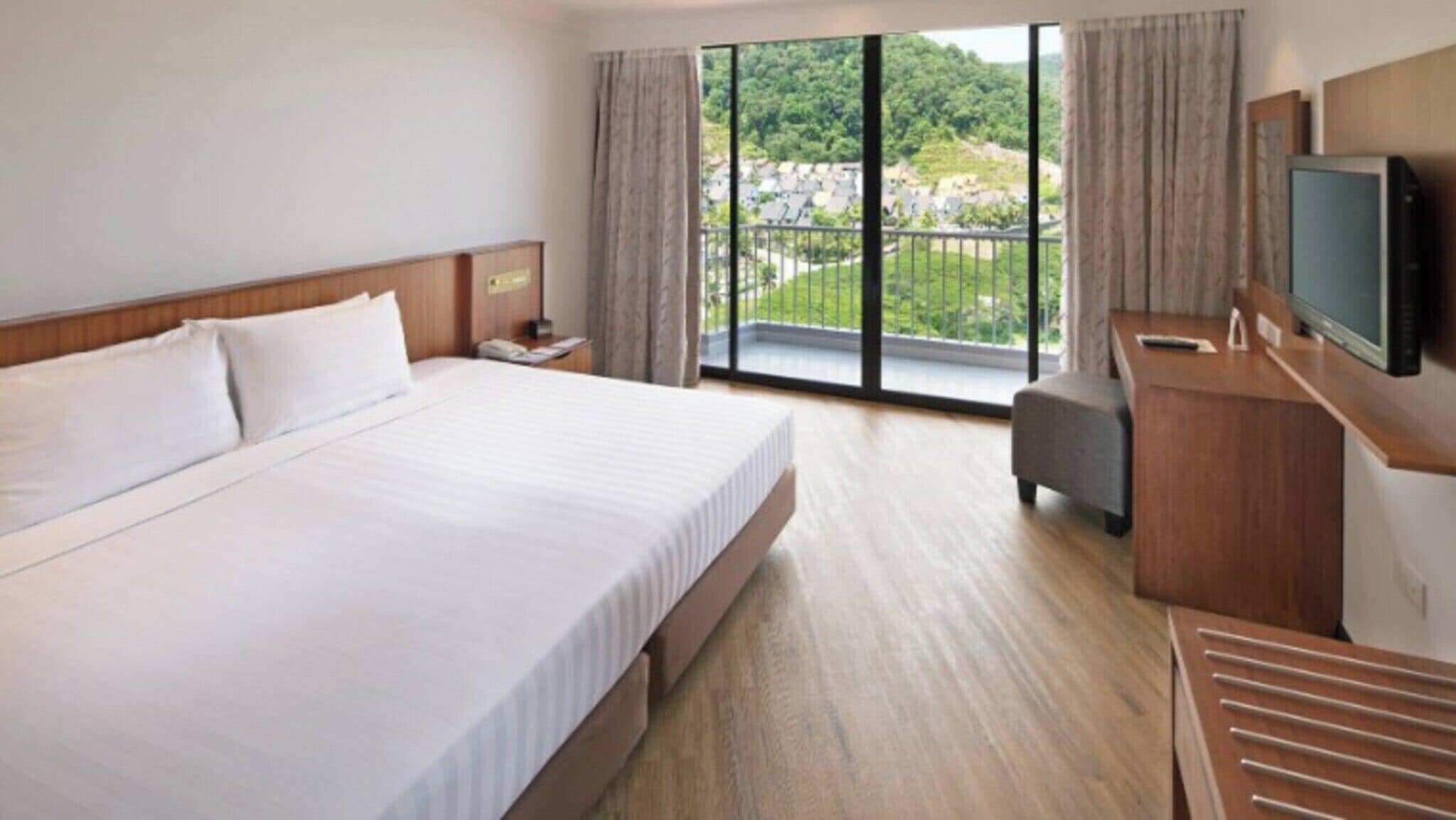 A hotel room with a large bed, a desk with a television, and a balcony with a view of greenery.