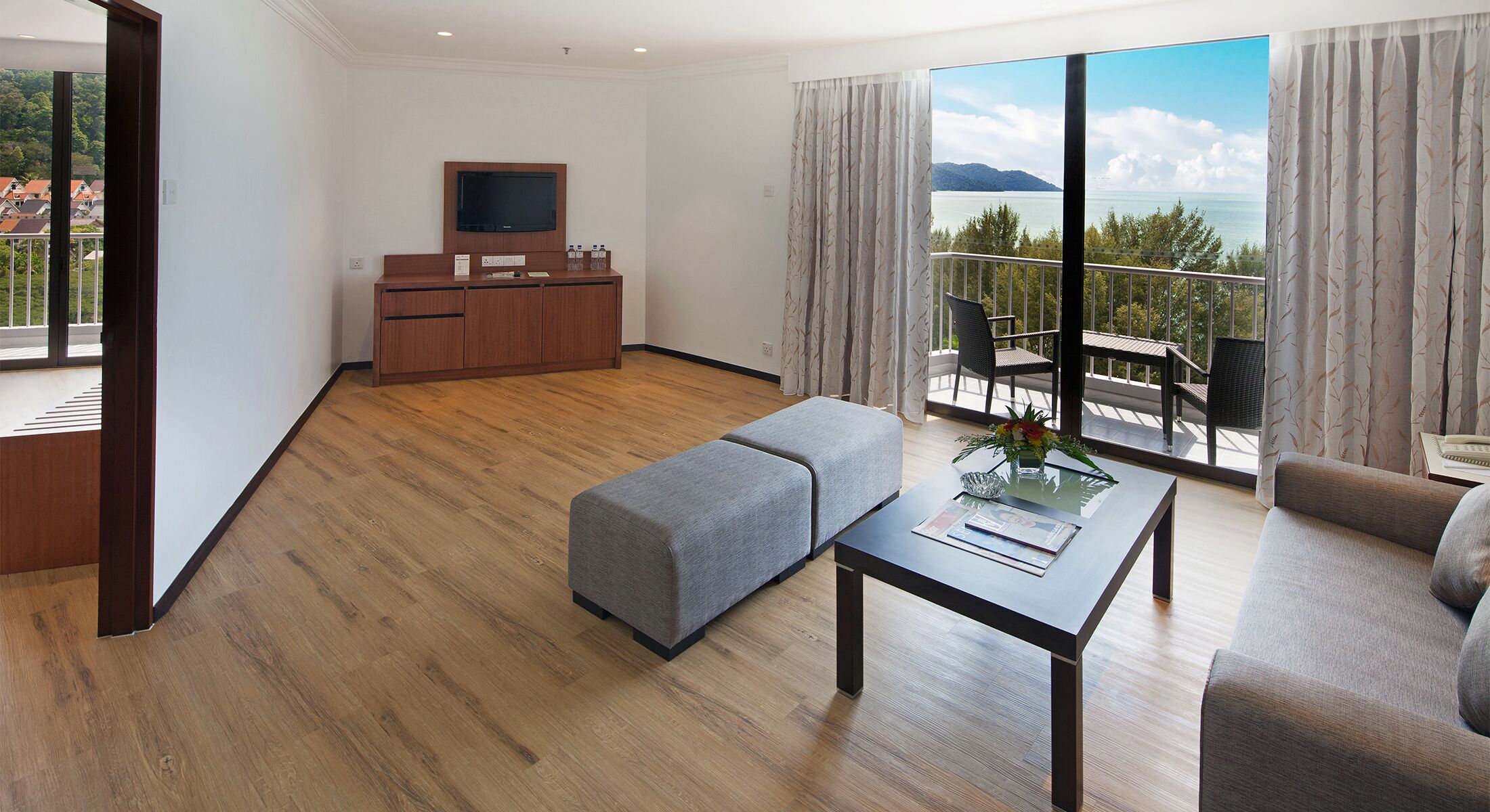 Een moderne hotelkamer met een houten vloer, een flatscreen televisie, een bank, een eettafel met een vaas en een balkon met uitzicht op zee.