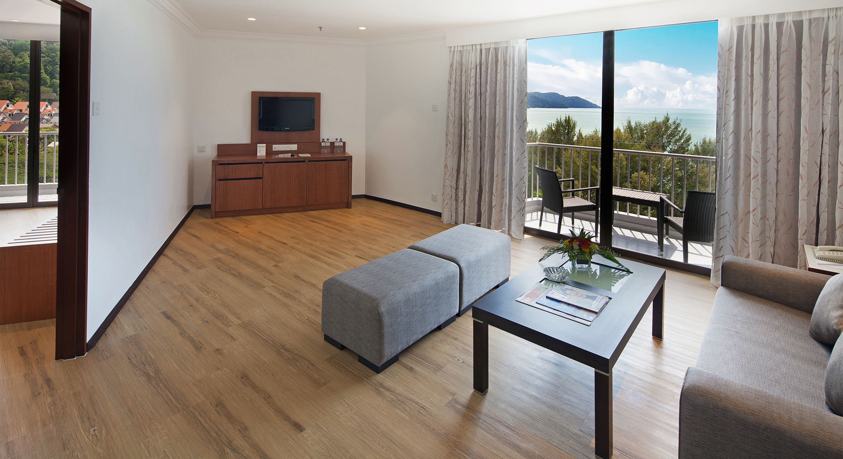 A modern hotel room with a wooden floor, a flat-screen TV, a sofa, a dining table, and a balcony with a view of the sea.