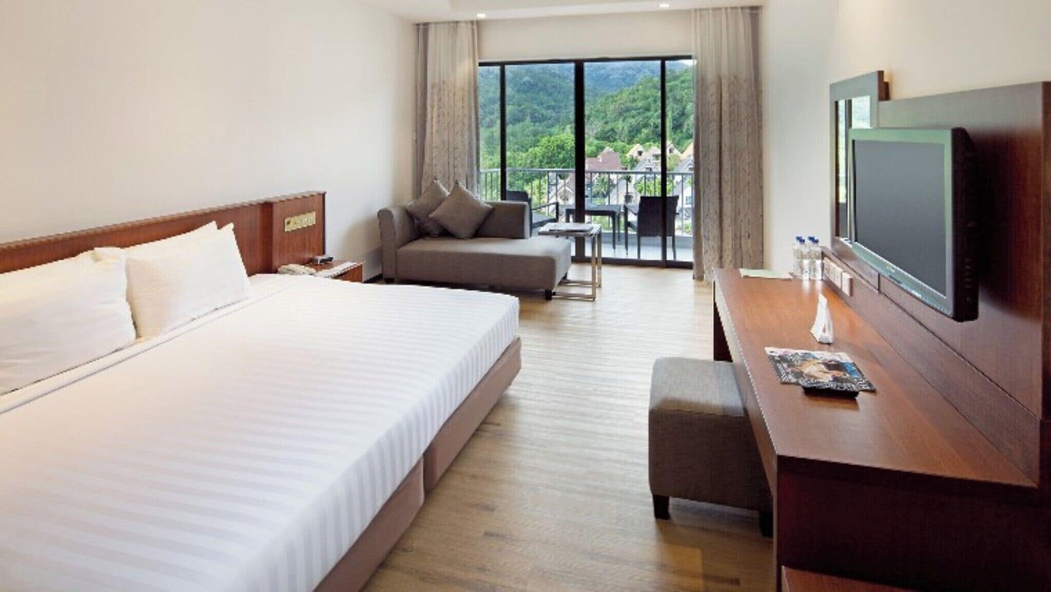 A hotel room with a large bed, a sofa, a desk with a TV, and a balcony with a view of greenery.