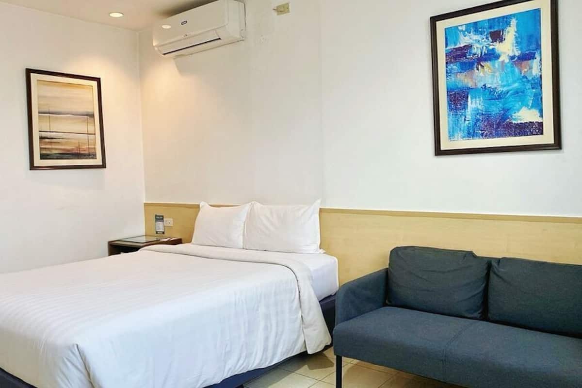 A hotel room with a large bed, a sofa, two framed artworks, and an air conditioning unit.