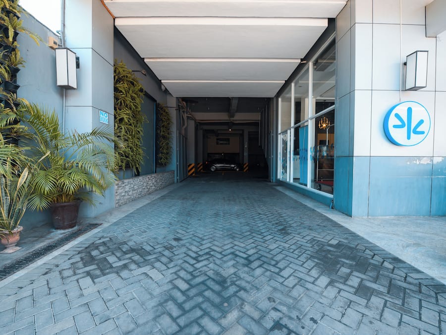 Un chemin pavé mène à l’entrée d’un bâtiment à la façade bleue et blanche, agrémenté de plantes en pot et d’un panneau indiquant une zone où le stationnement est interdit.