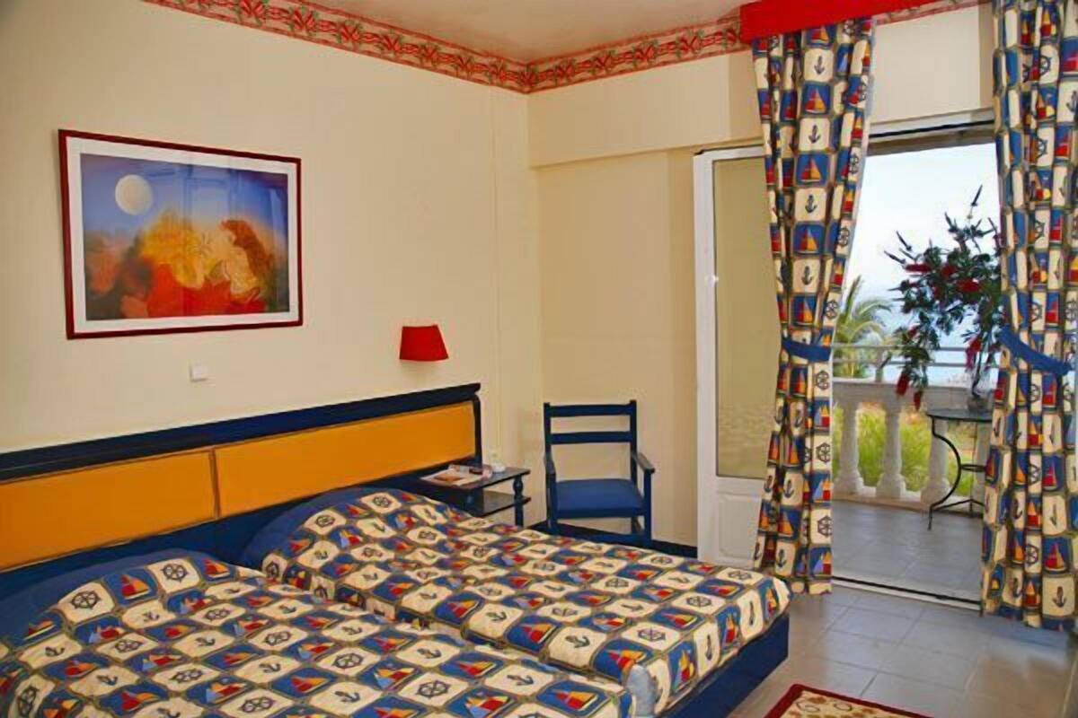 A bedroom with a bed, a chair, a painting, and a balcony with patterned curtains.